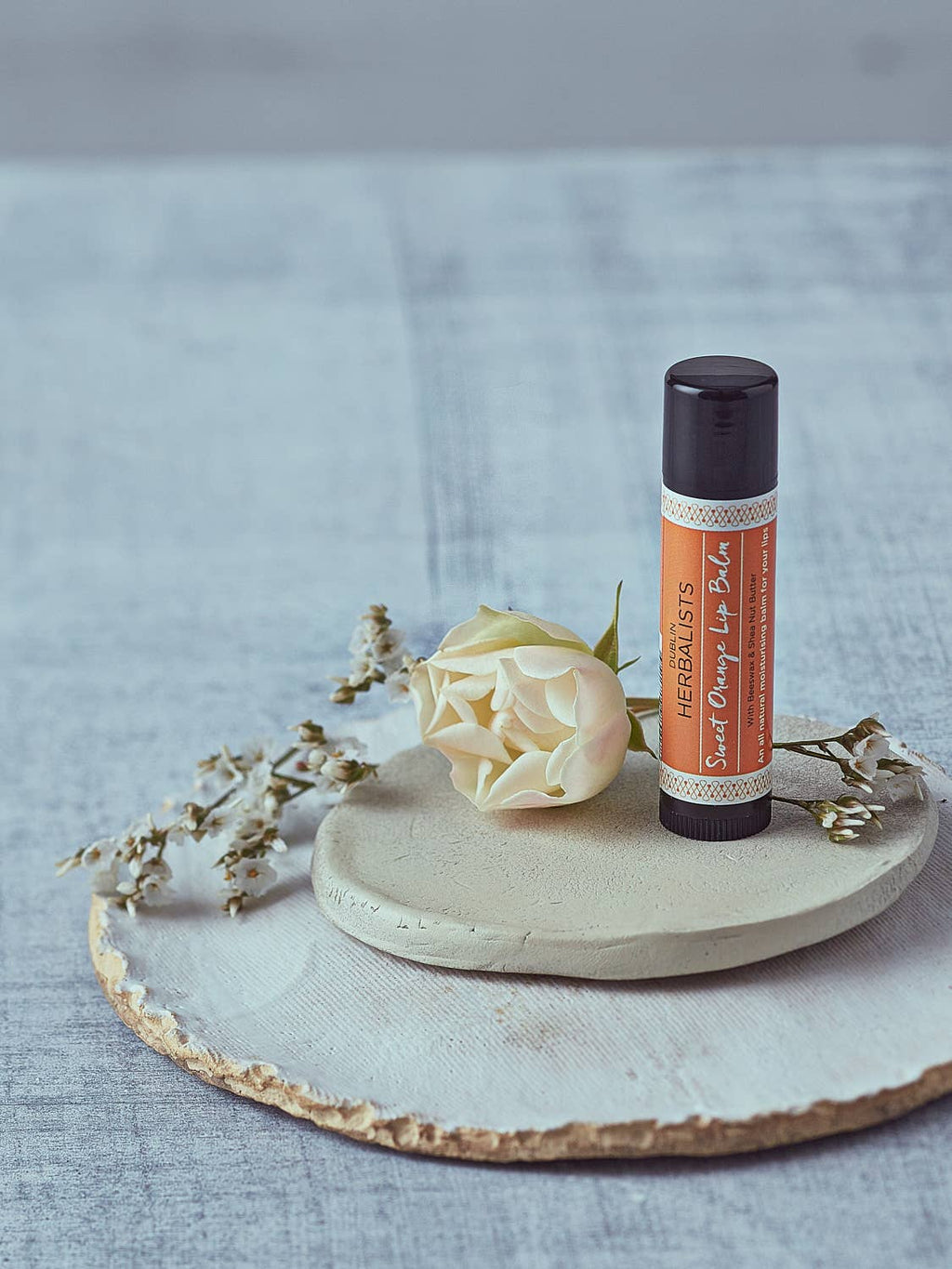 Sweet Orange Lip Balm tube standing on a ceramic dish with a white rose